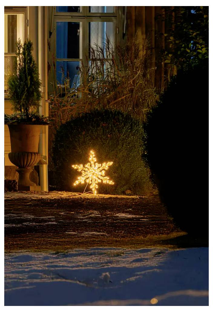 Une décoration lumineuse en forme de flocon de neige brillant brille dans une cour avant enneigée à côté d'une maison la nuit.