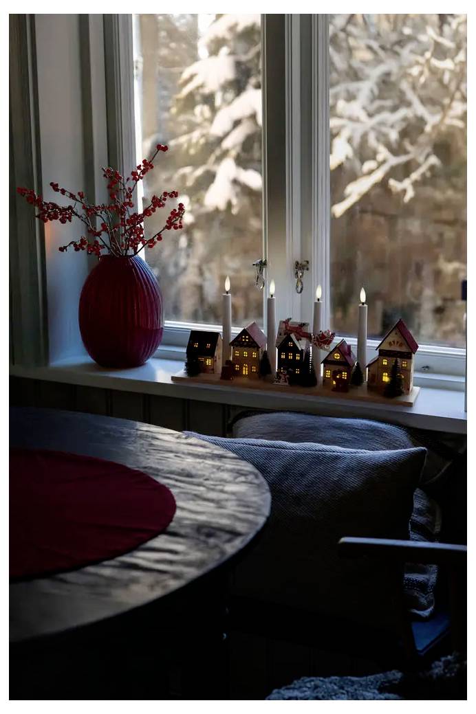 Rebord de fenêtre douillet d'hiver avec un vase rouge rempli de baies et de petites maisons miniatures illuminées, aperçu à côté d'une table et d'une chaise avec des arbres enneigés à l'extérieur.