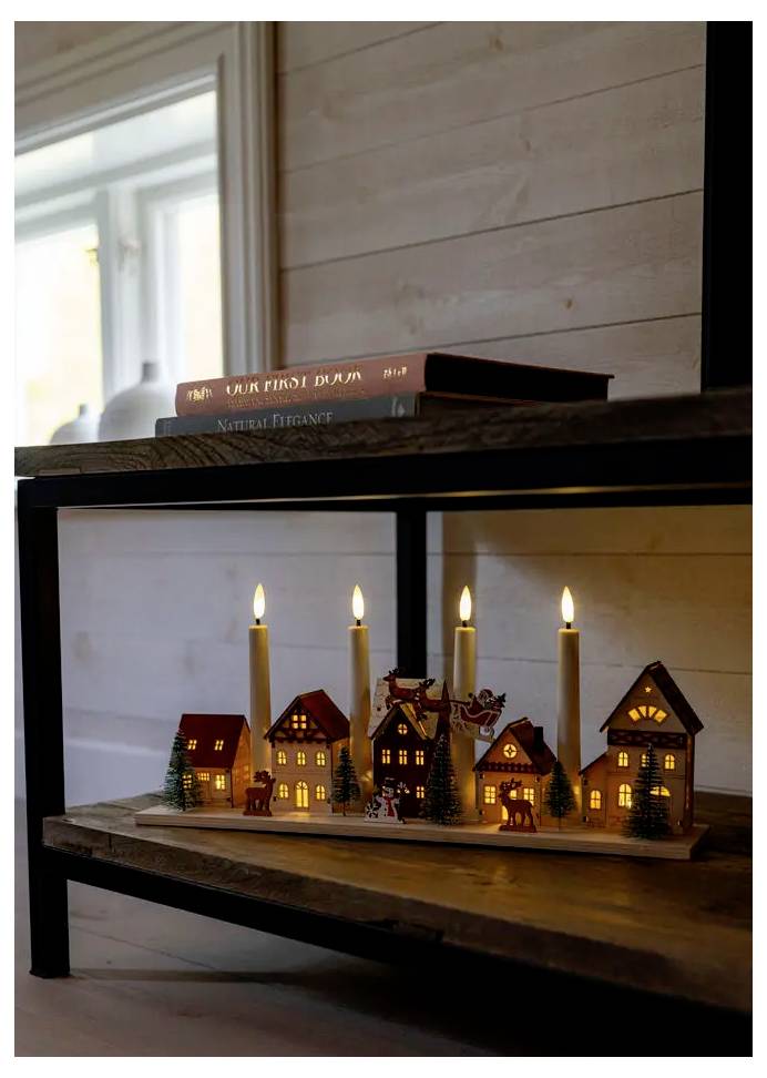 Des petites maisons de village miniatures illuminées par des bougies sur une étagère en bois sous deux livres empilés dans une pièce intérieure douillette.