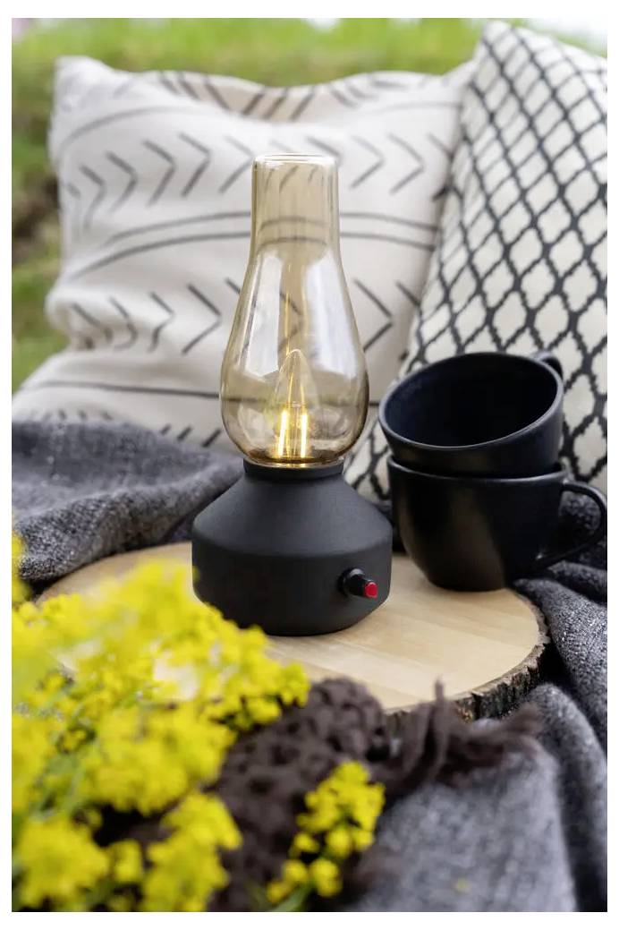 Scène de table avec une lampe à bougie en verre allumée sur une base noire, des tasses noires, des coussins et des fleurs jaunes créant un cadre extérieur douillet.