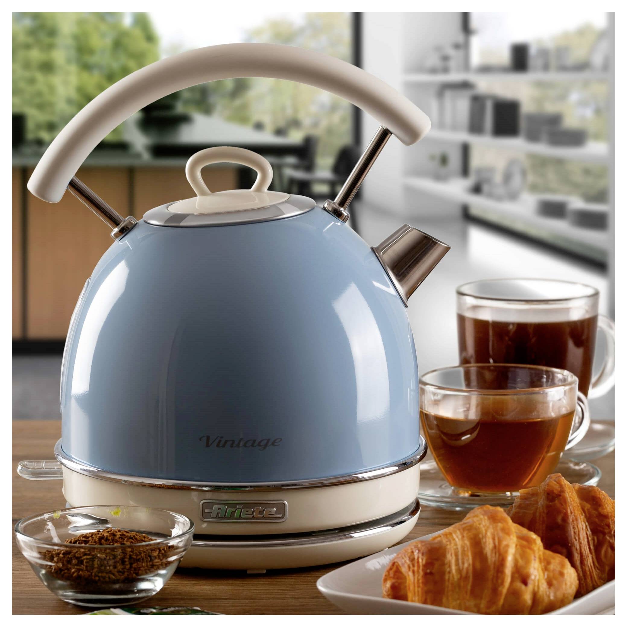 Bouilloire électrique bleue sur une table avec du thé et des croissants, dans un environnement de cuisine lumineux.