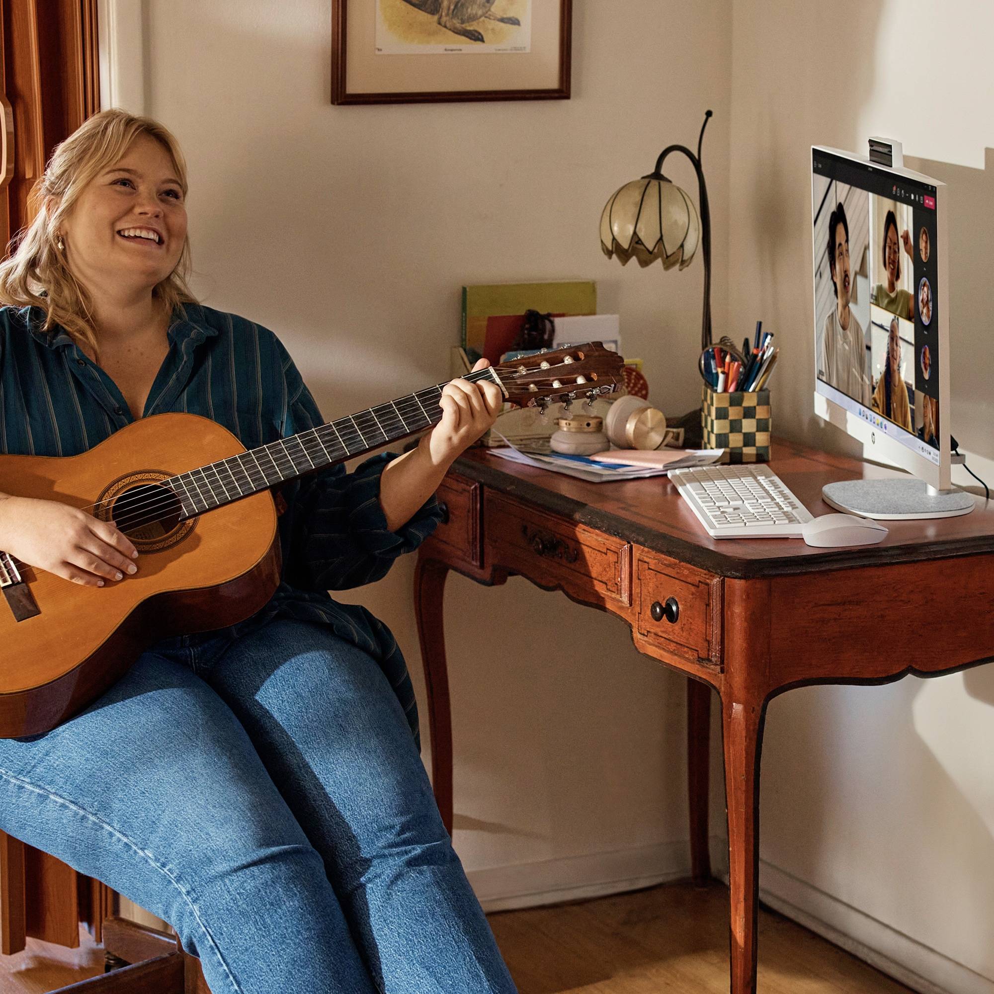 Femme souriante jouant de la guitare acoustique à un bureau lors d'un appel vidéo sur un écran d'ordinateur.