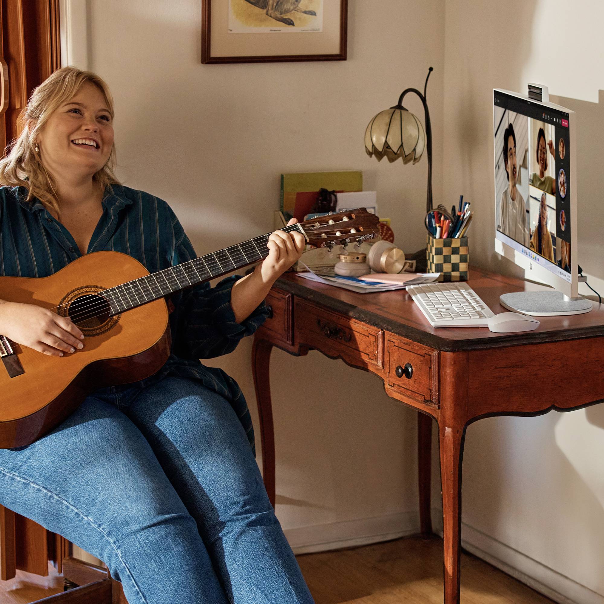 Femme jouant de la guitare lors d'un appel vidéo à un bureau à domicile, avec un ordinateur affichant plusieurs participants à l'écran.