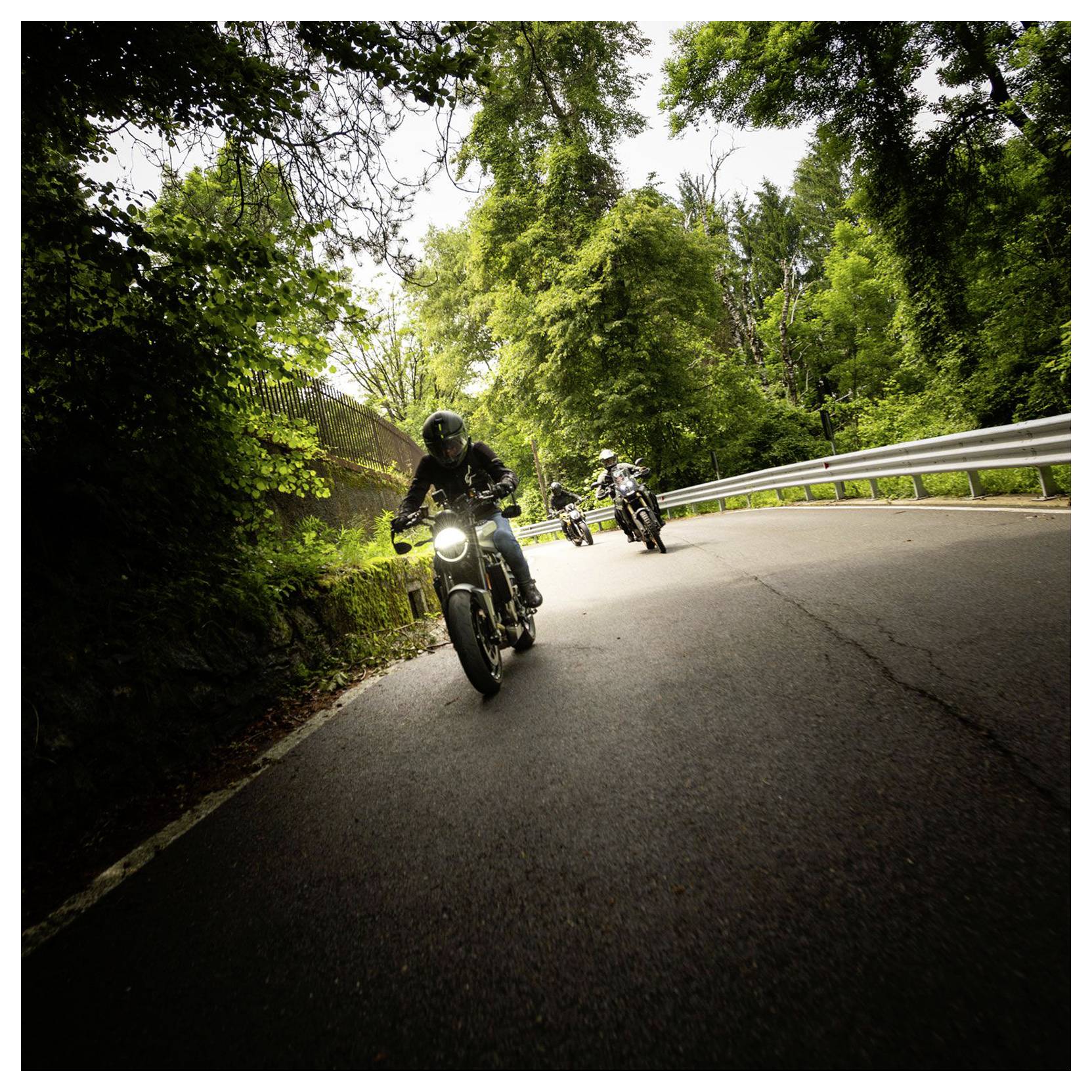 Trois motocyclistes roulent sur une route sinueuse bordée d'arbres, la moto de tête se dirigeant vers la caméra avec son phare allumé.