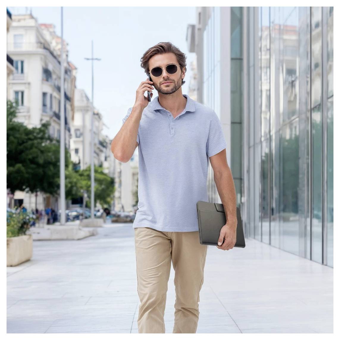 Un homme marchant sur un trottoir de la ville en parlant au téléphone et portant une chemise à dossiers pour ordinateur portable, portant des lunettes de soleil et un polo bleu clair.