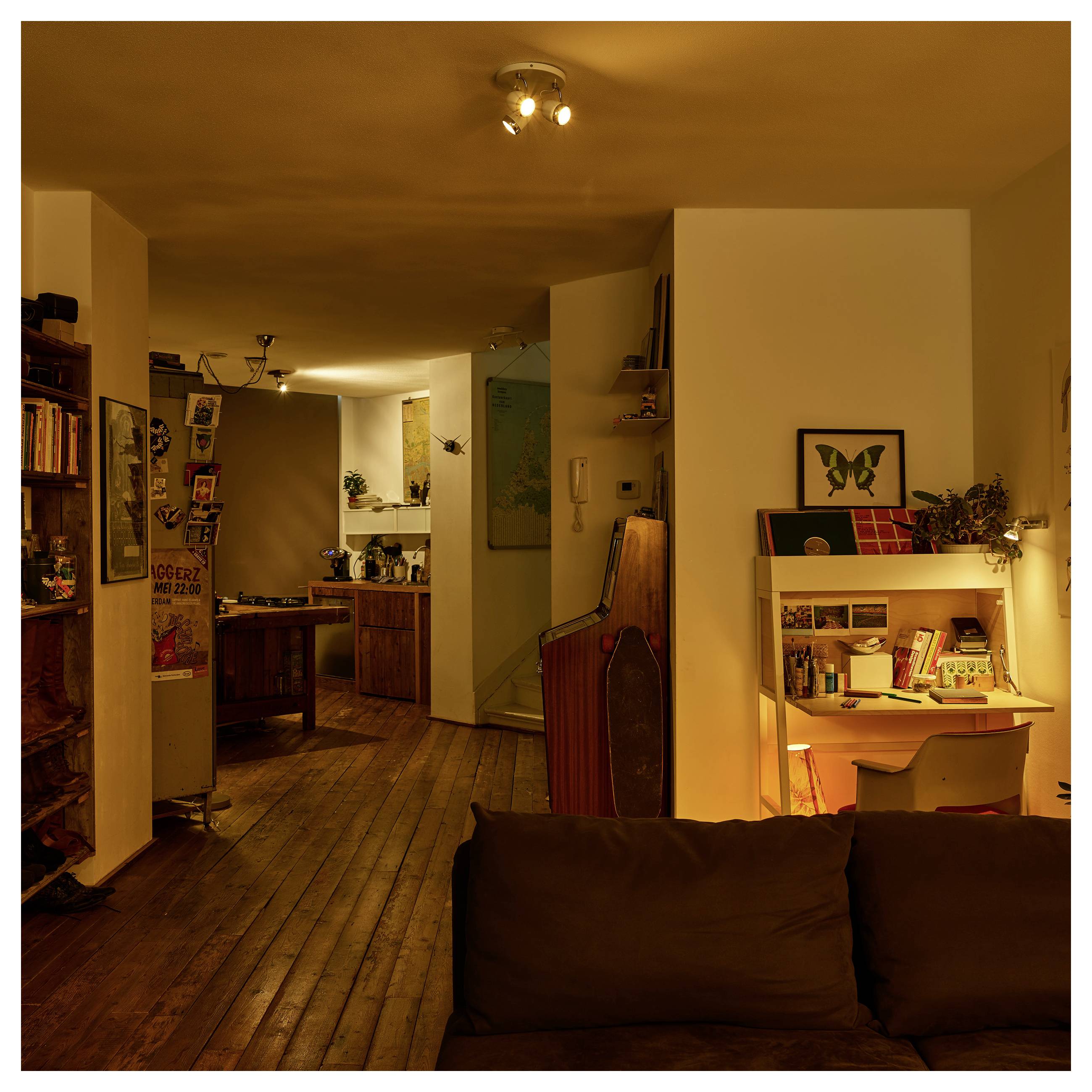 Intérieur d'appartement chaleureusement éclairé avec des planchers en bois, un canapé foncé au premier plan, un petit coin bureau à droite et une cuisine en arrière-plan.