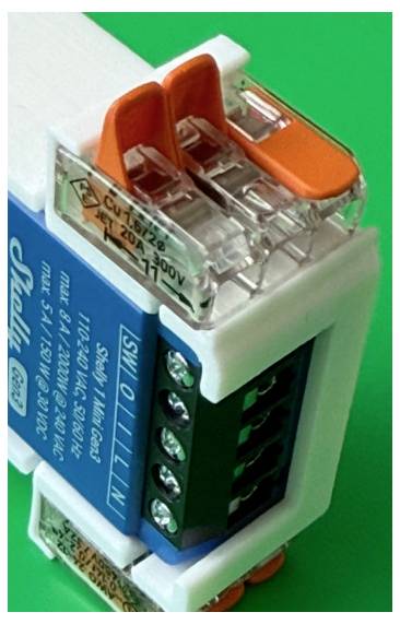 Gros plan d'un module de relais électrique avec couvercle transparent et pinces orange, montrant les bornes à vis et une étiquette bleue sur un fond vert.
