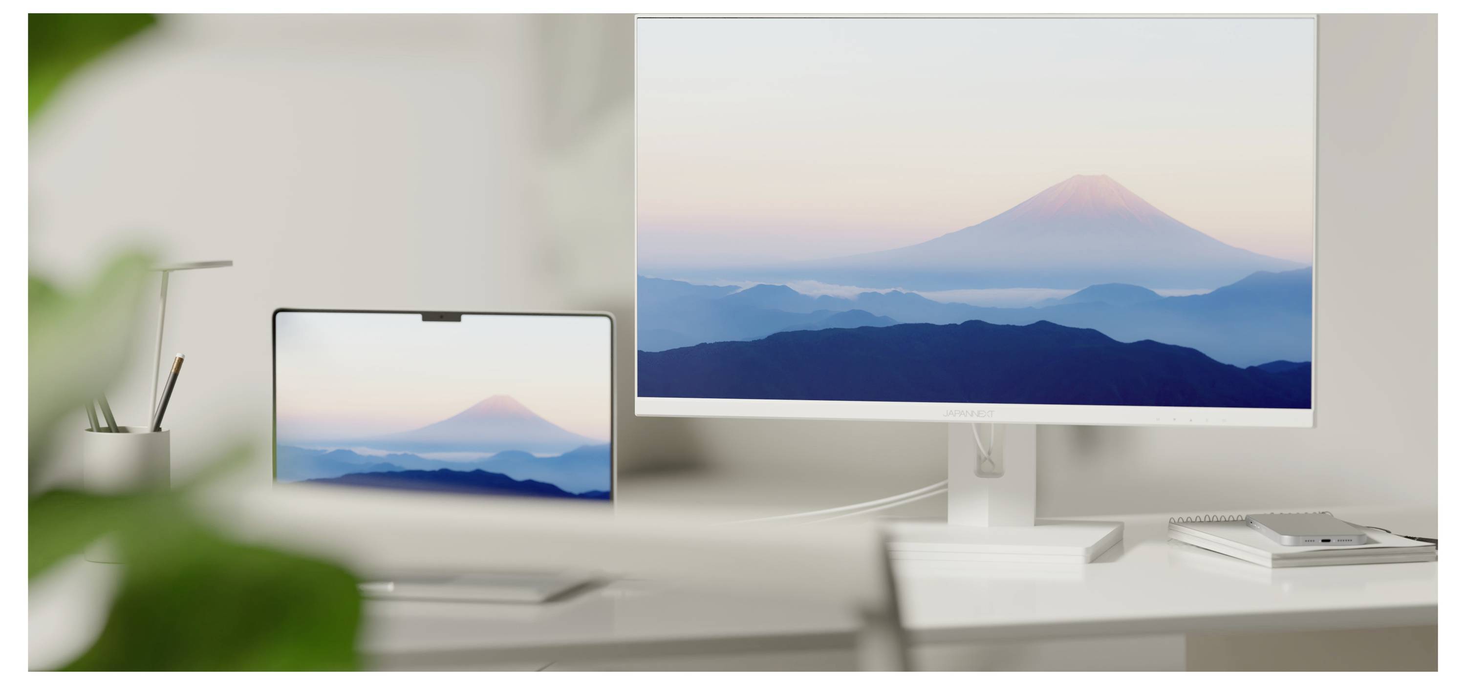 Un bureau aménagé avec un moniteur et une tablette affichant tous deux une montagne brumeuse au lever du soleil, vue depuis un espace de travail propre.