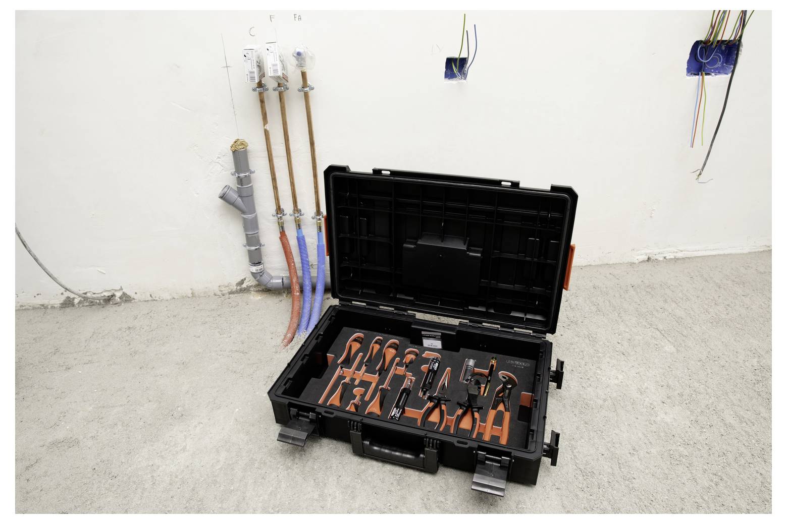 Mallette noire ouverte avec des outils manuels à poignées orange sur un sol en béton, à côté de tuyauterie murale et de câblage électrique dans une pièce inachevée.