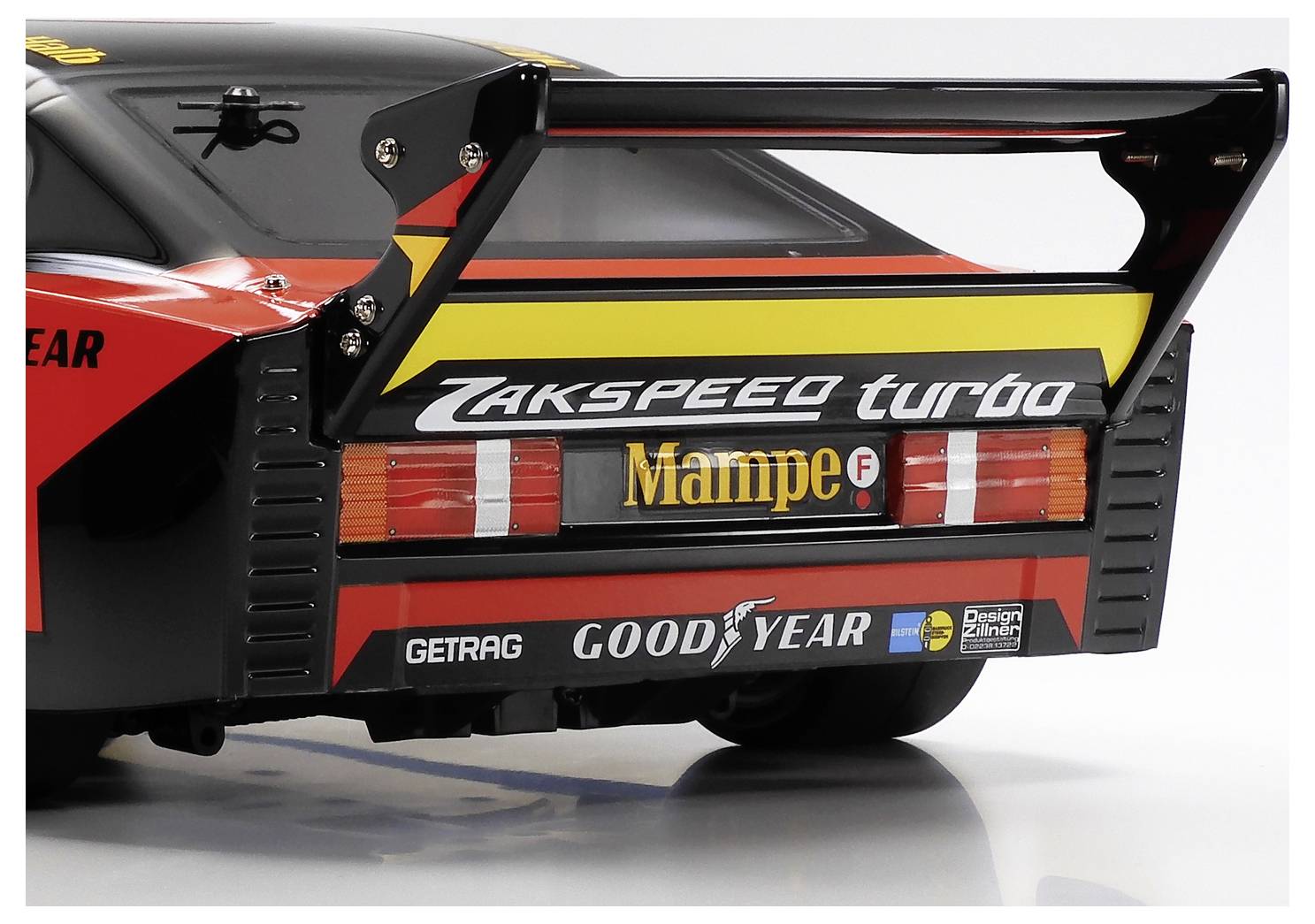 Vue arrière d'une voiture de course noire et rouge montrant un grand aileron arrière et des autocollants de parrainage incluant 'Zakspeed turbo', 'Mampe', 'Goodyear', et 'GETRAG'.