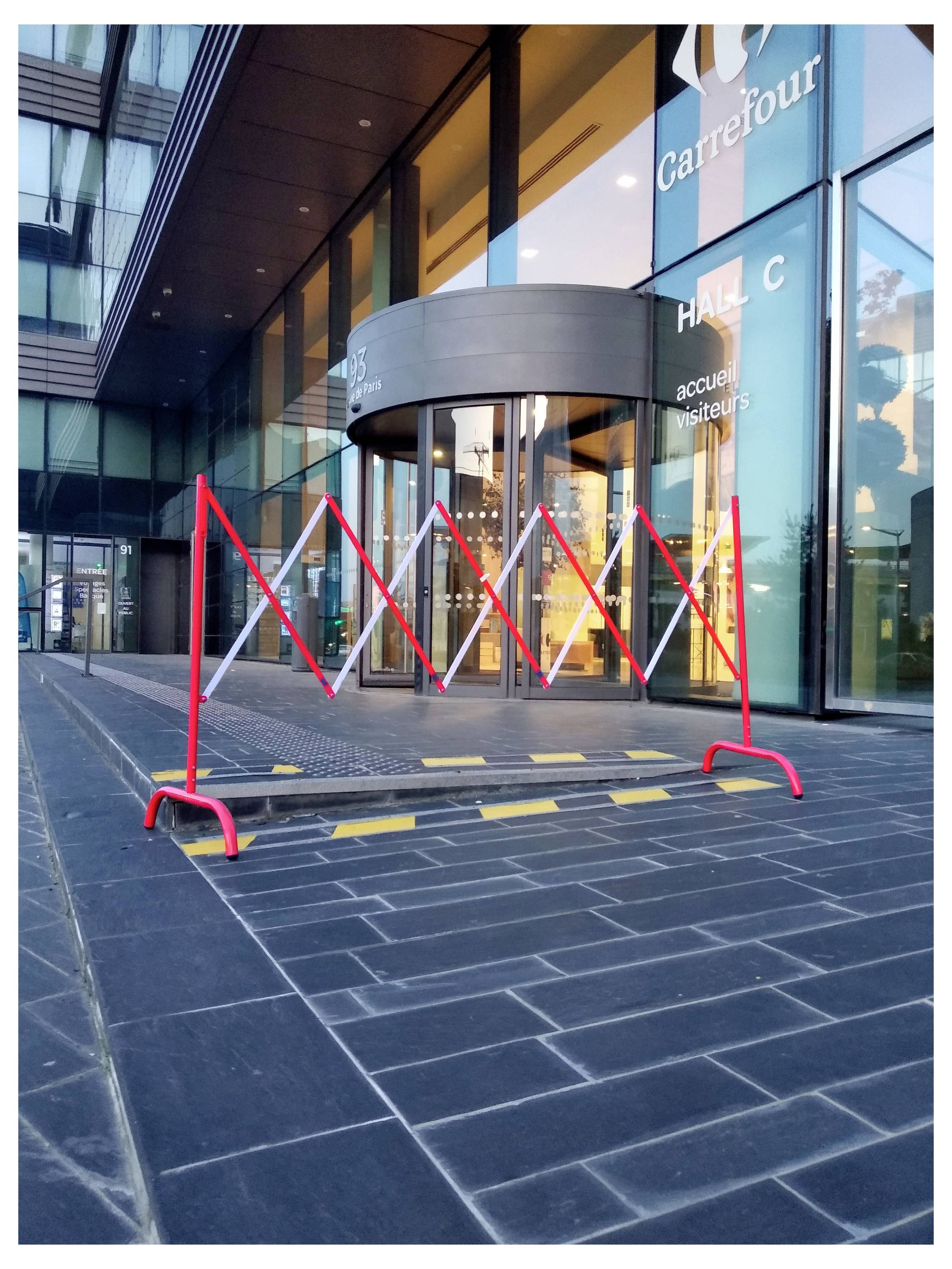 Une barrière temporaire rouge et blanche bloque l'entrée d'un bâtiment Carrefour étiqueté « HALL C » et « accueil visiteurs ».