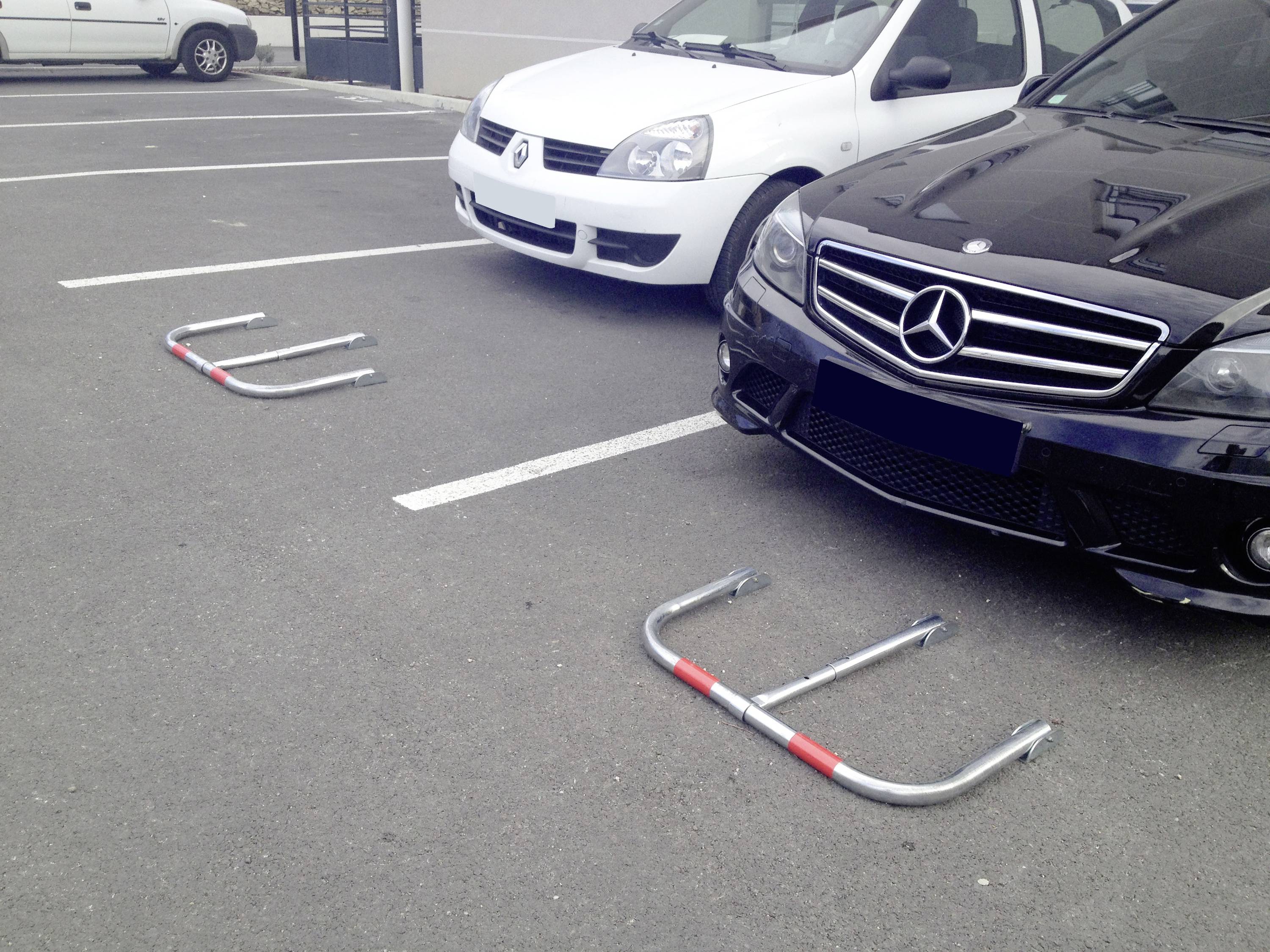 Deux sabots de roue sont posés sur le trottoir devant des voitures garées, dont une Mercedes noire et une Renault blanche, dans un parking.