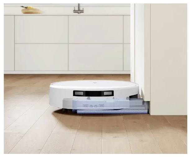 Un robot aspirateur nettoyant un sol de cuisine en bois tandis qu'il se positionne à côté d'un meuble dans une cuisine moderne de couleur blanche.