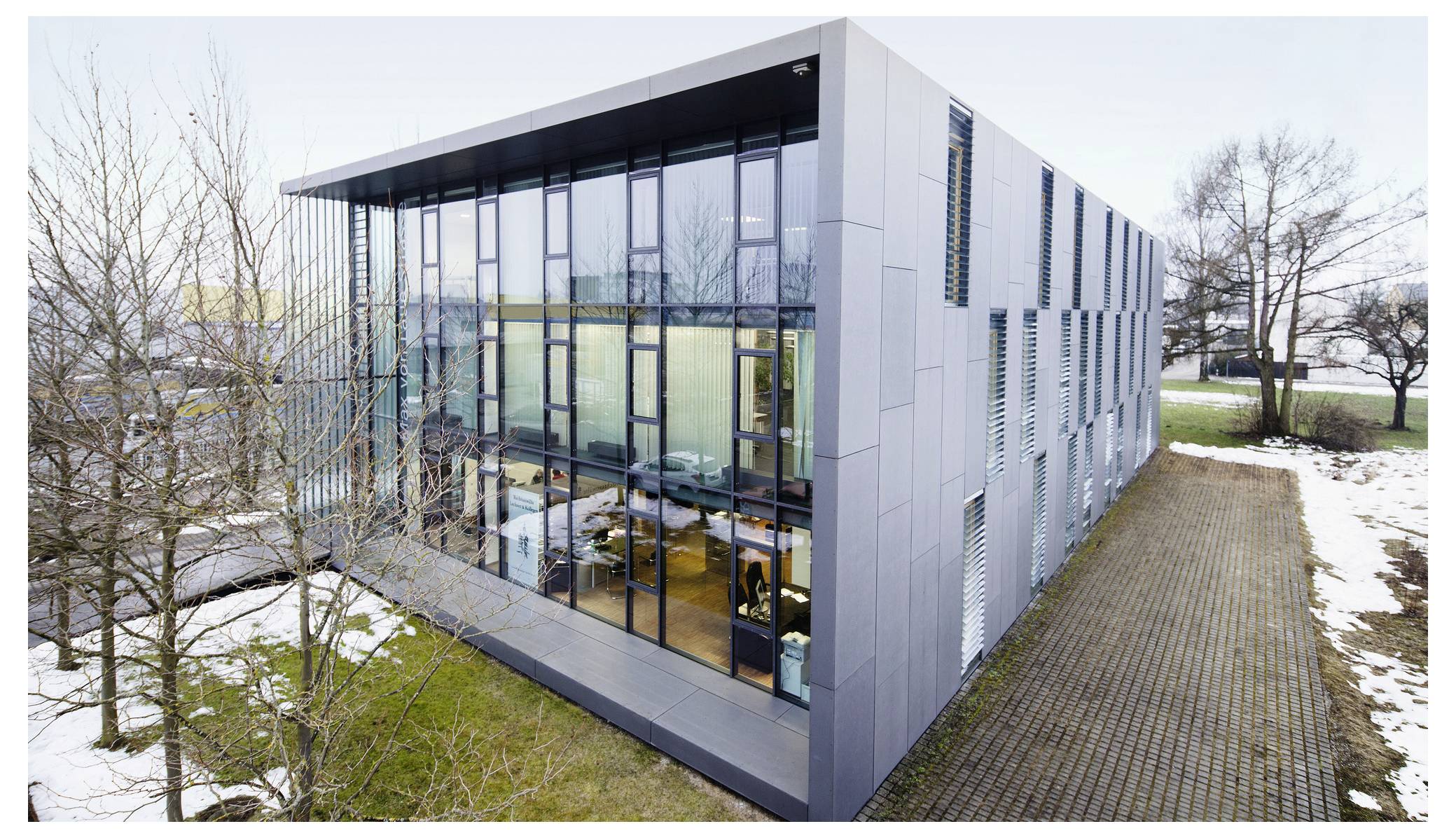Bâtiment de bureaux moderne avec un coin à façade vitrée, vu de dessus, à côté d'un sentier pavé et d'arbres épars en hiver avec des plaques de neige.