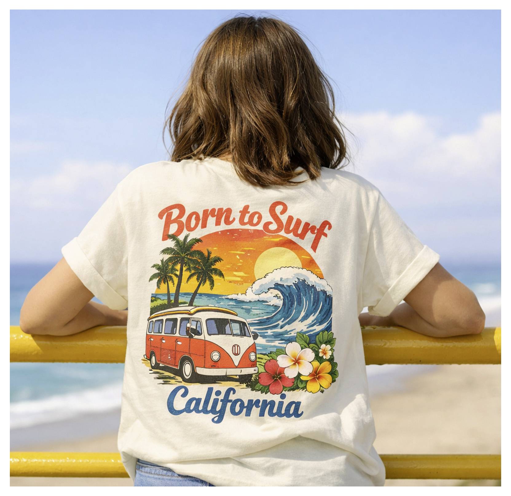 Femme vue de dos portant un t-shirt « Born to Surf California » avec un graphique représentant un van de plage, une vague et un palmier, debout près d'une barrière en bord de mer.