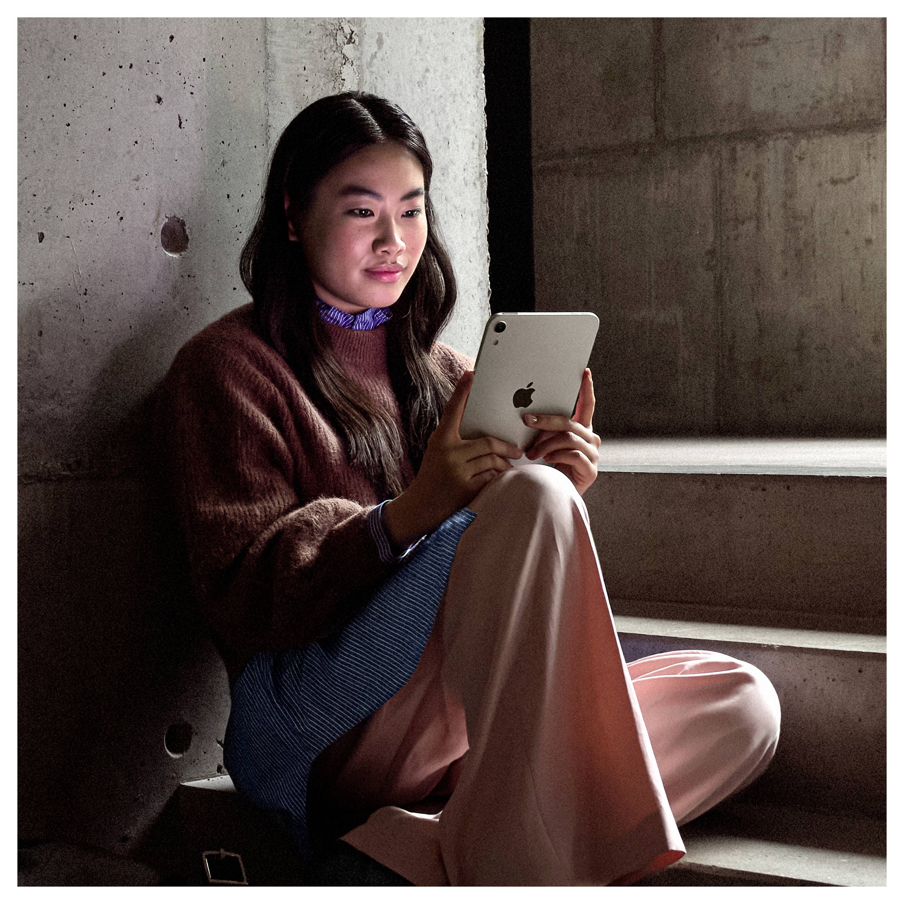 Jeune femme assise sur des marches en béton, regardant et utilisant une tablette dans un espace intérieur faiblement éclairé.