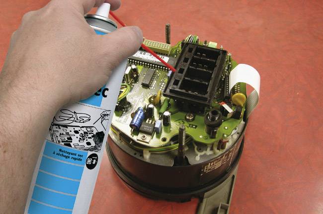 La main pulvérise un produit de nettoyage sur un circuit électronique avec carte de circuit imprimé, résistant à la poussière avec un aérosol sur un fond rouge.