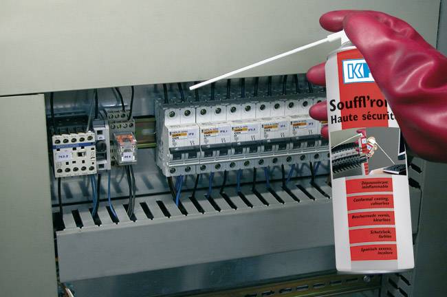 Un électricien utilise un aérosol à air comprimé dans un tableau de distribution pour enlever la poussière. Il porte des gants de protection rouges.