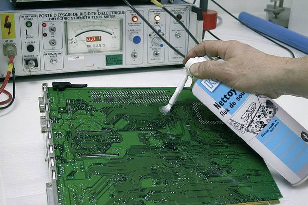 Une main nettoie une carte électronique verte avec un spray et un coton-tige. En arrière-plan, un appareil de test avec différents affichages est visible.