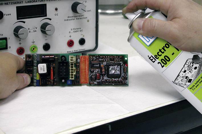 Une personne nettoie une carte électronique avec un spray, à côté d'un appareil de laboratoire équipé de divers boutons rotatifs et affichages.