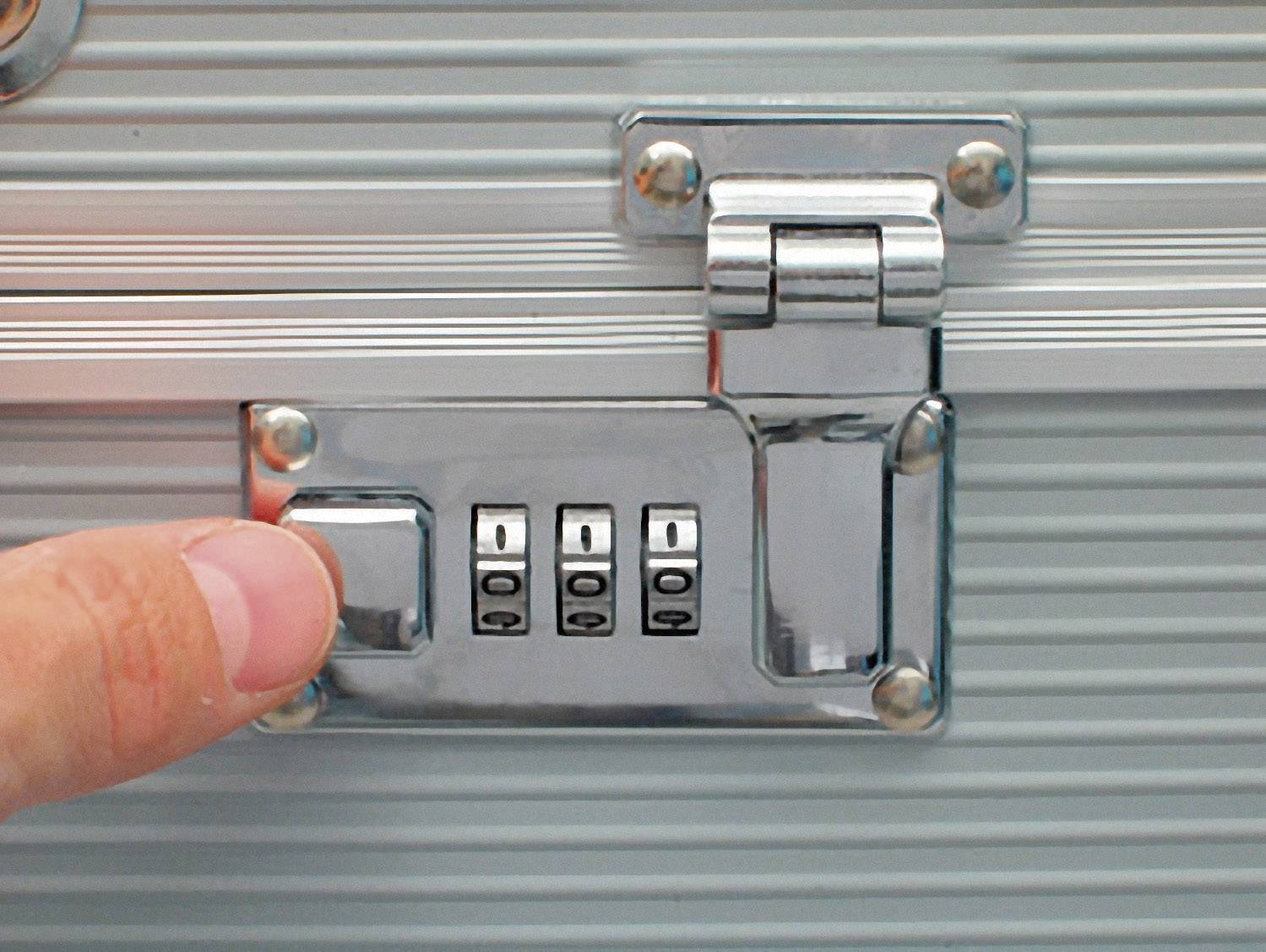 Une main appuie sur le levier d'un cadenas à combinaison sur une valise argentée.