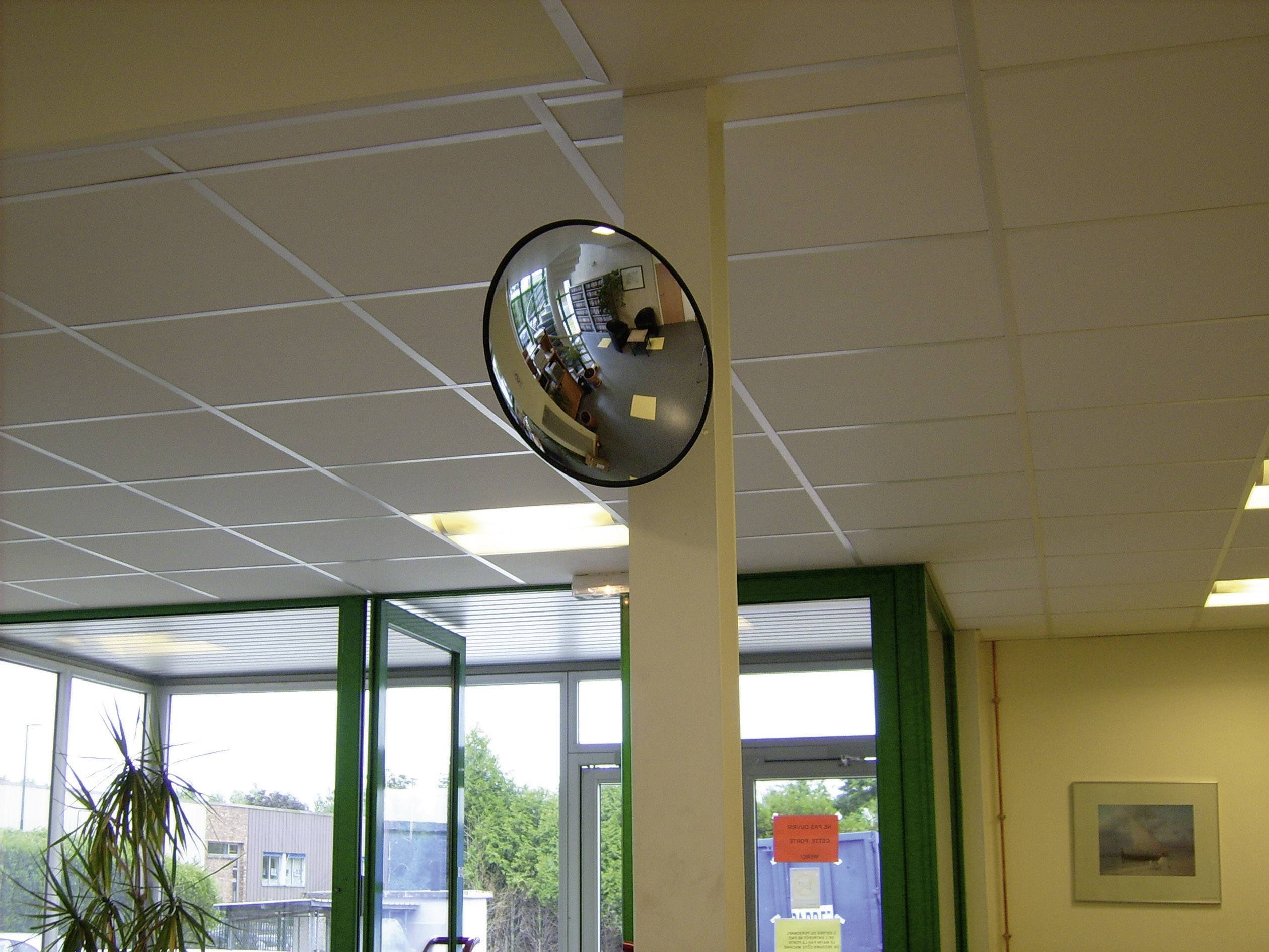 Un miroir rond et bombé est suspendu dans un coin d'un intérieur, reflétant les meubles et les fenêtres, offrant une vue d'ensemble de la pièce.