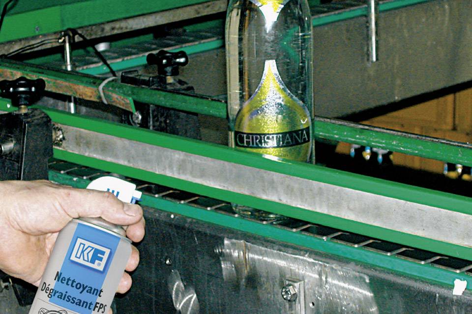 Une personne tient une bombe aérosol 'Nettoyant Dégraissant FP'. En arrière-plan, une bouteille 'Christiana' est sur un tapis roulant dans une ligne de production.