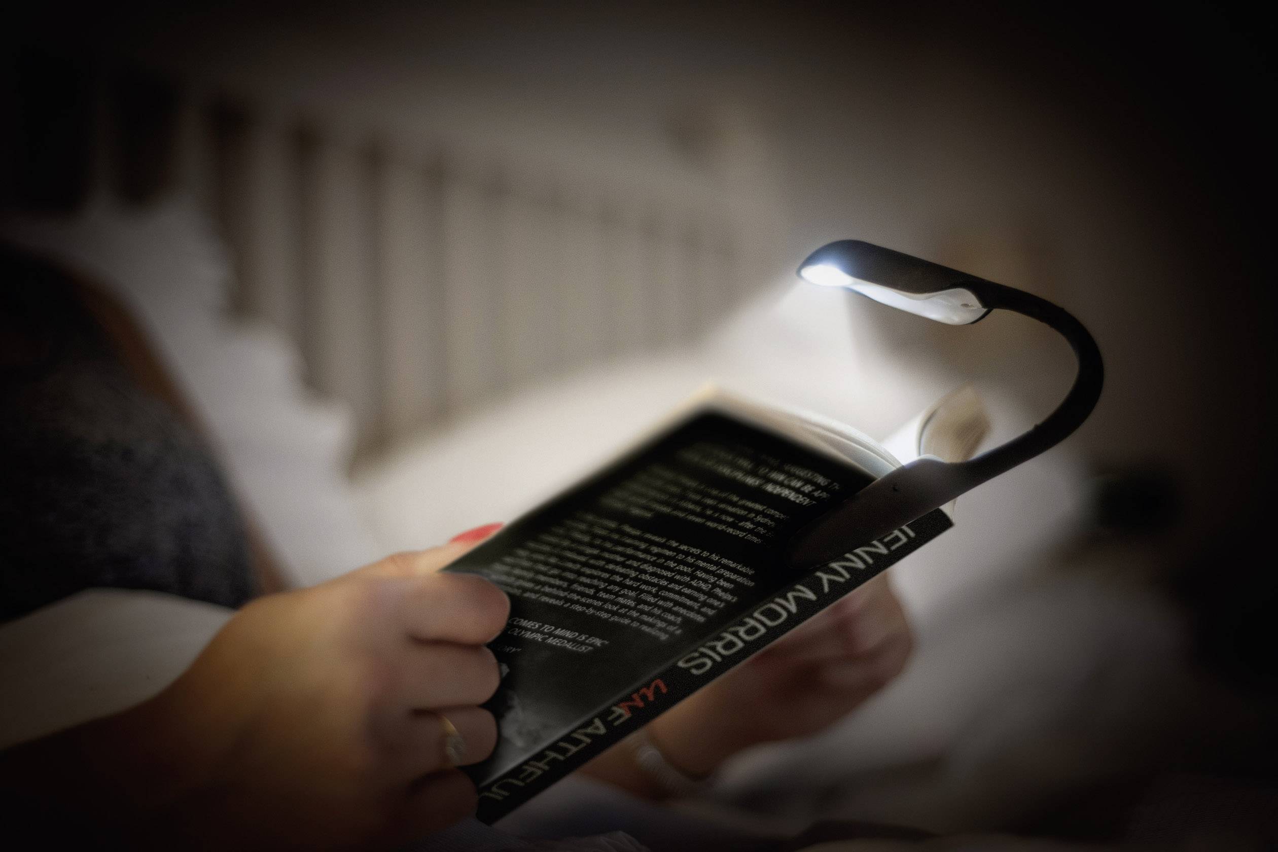Une personne lit la nuit dans son lit à l'aide d'une lampe de lecture dirigée vers le livre. La chambre est faiblement éclairée.