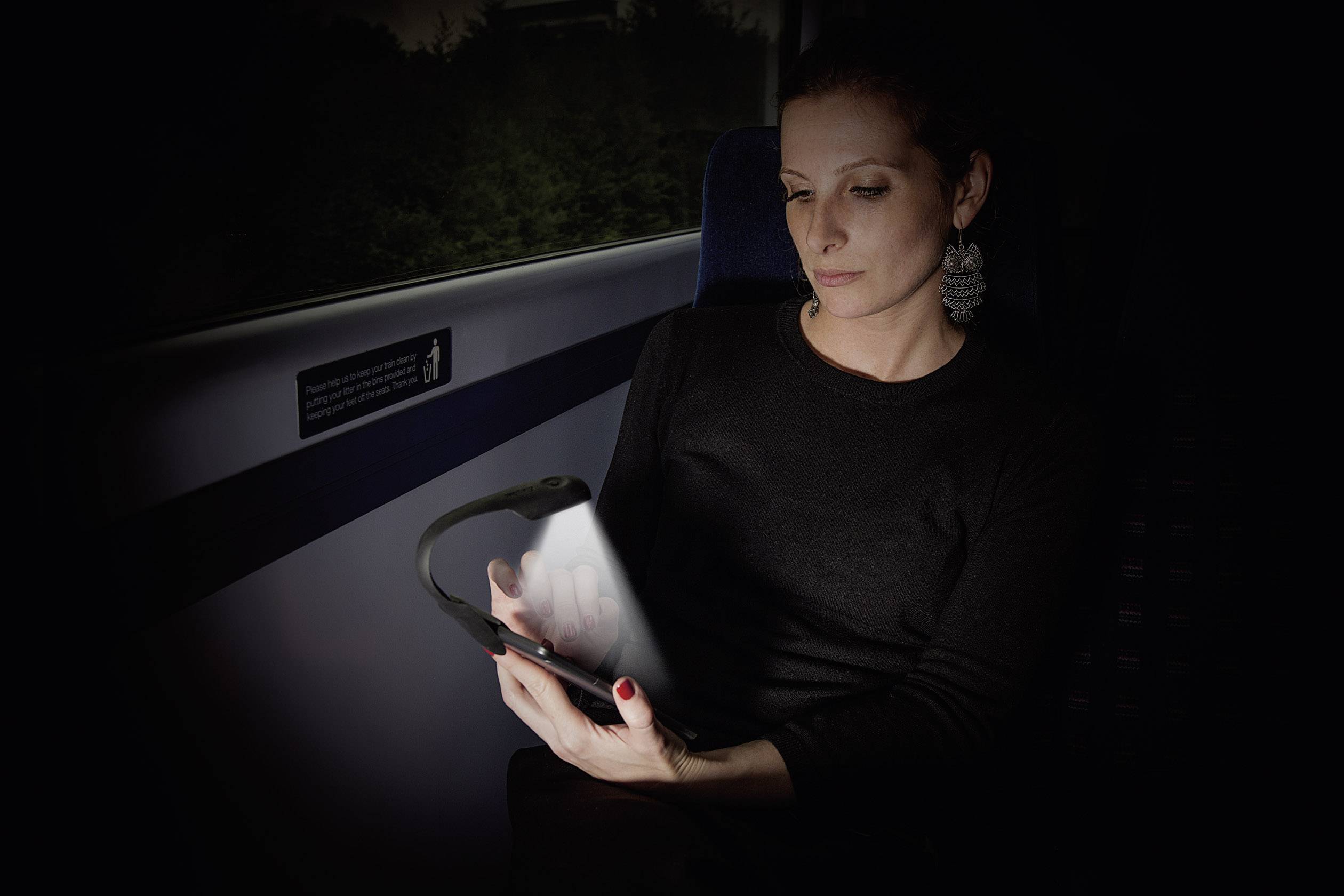 Une femme est assise dans un compartiment de train sombre et lit sur une tablette éclairée. Elle porte des vêtements noirs et des boucles d'oreilles.