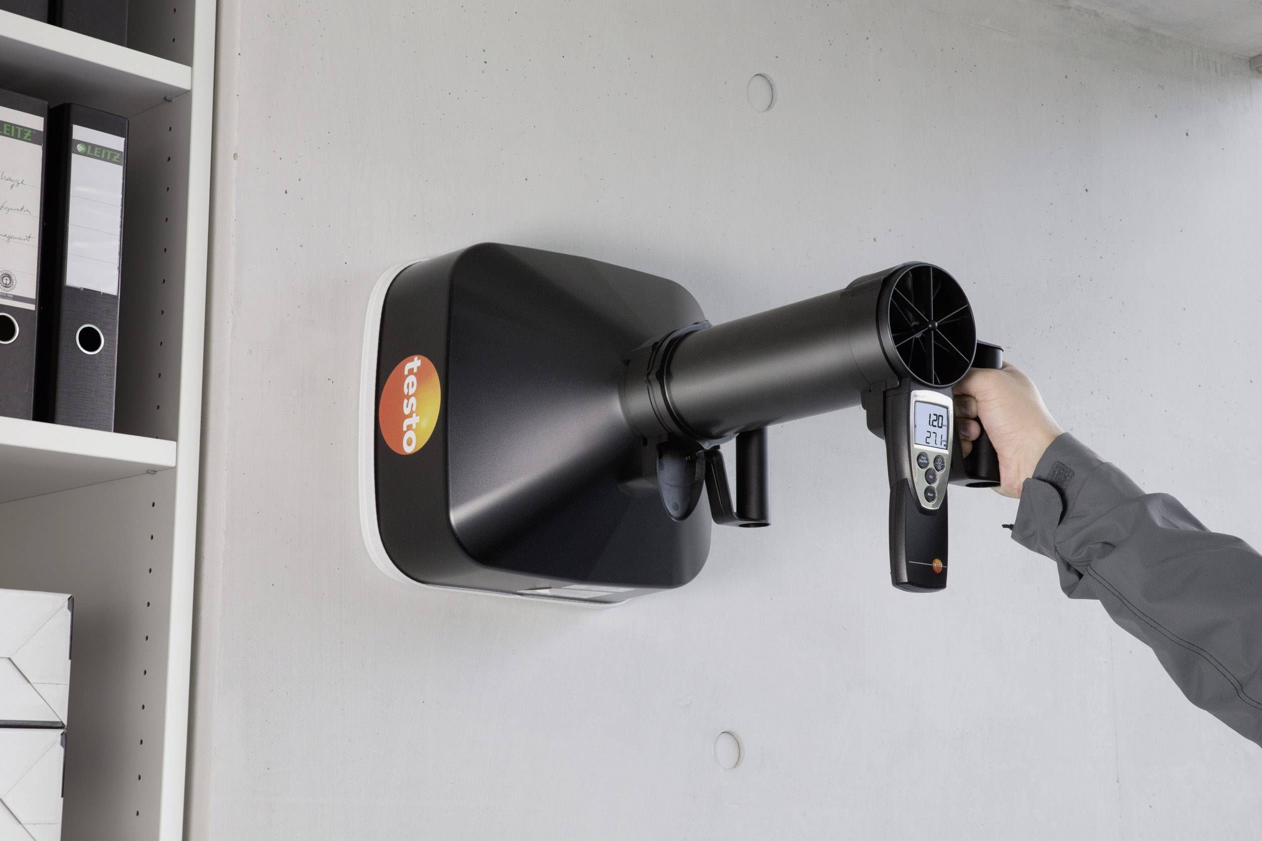 Une personne utilise un appareil de mesure de ventilation monté sur le mur pour tester le débit d'air. Des dossiers sont visibles à gauche de l'image.