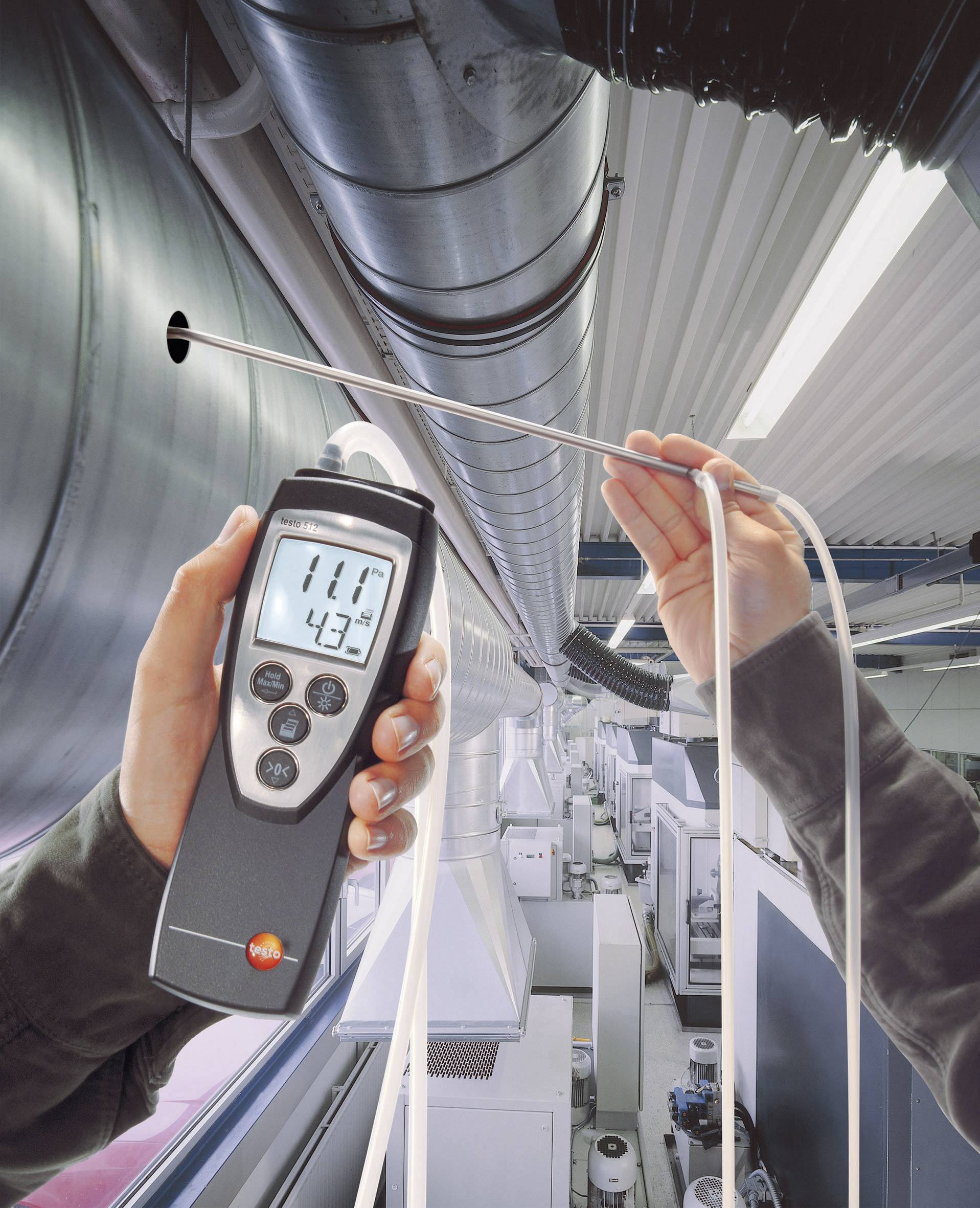 Une personne mesure la température de l'air et l'humidité dans un système de ventilation industriel à l'aide d'un appareil portable. L'écran affiche '11°C' et '43%'.