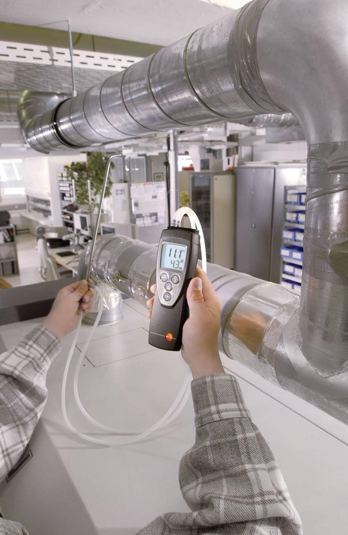 Une personne mesure la qualité de l'air dans un bureau à l'aide d'un appareil de mesure numérique. L'écran affiche une valeur de 119. Des conduits de ventilation sont visibles en arrière-plan.