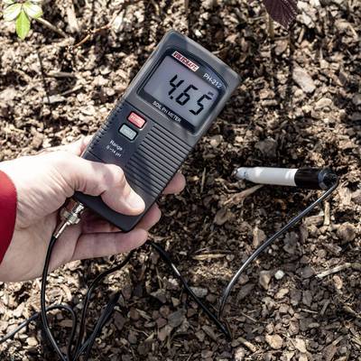 Une personne tient un pH-mètre numérique au-dessus de la terre, affichant une valeur de pH de 4,05. Le capteur est enfoncé dans le sol.