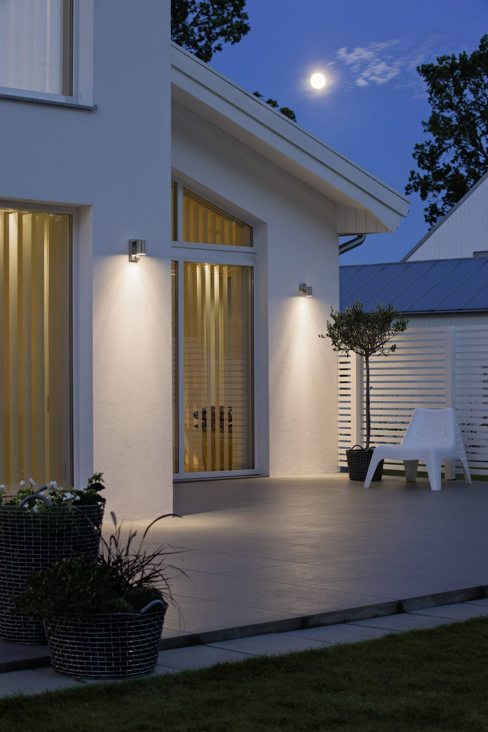 Une maison moderne la nuit avec une terrasse éclairée, la lune dans le ciel, des plantes en pot et une chaise blanche au premier plan.