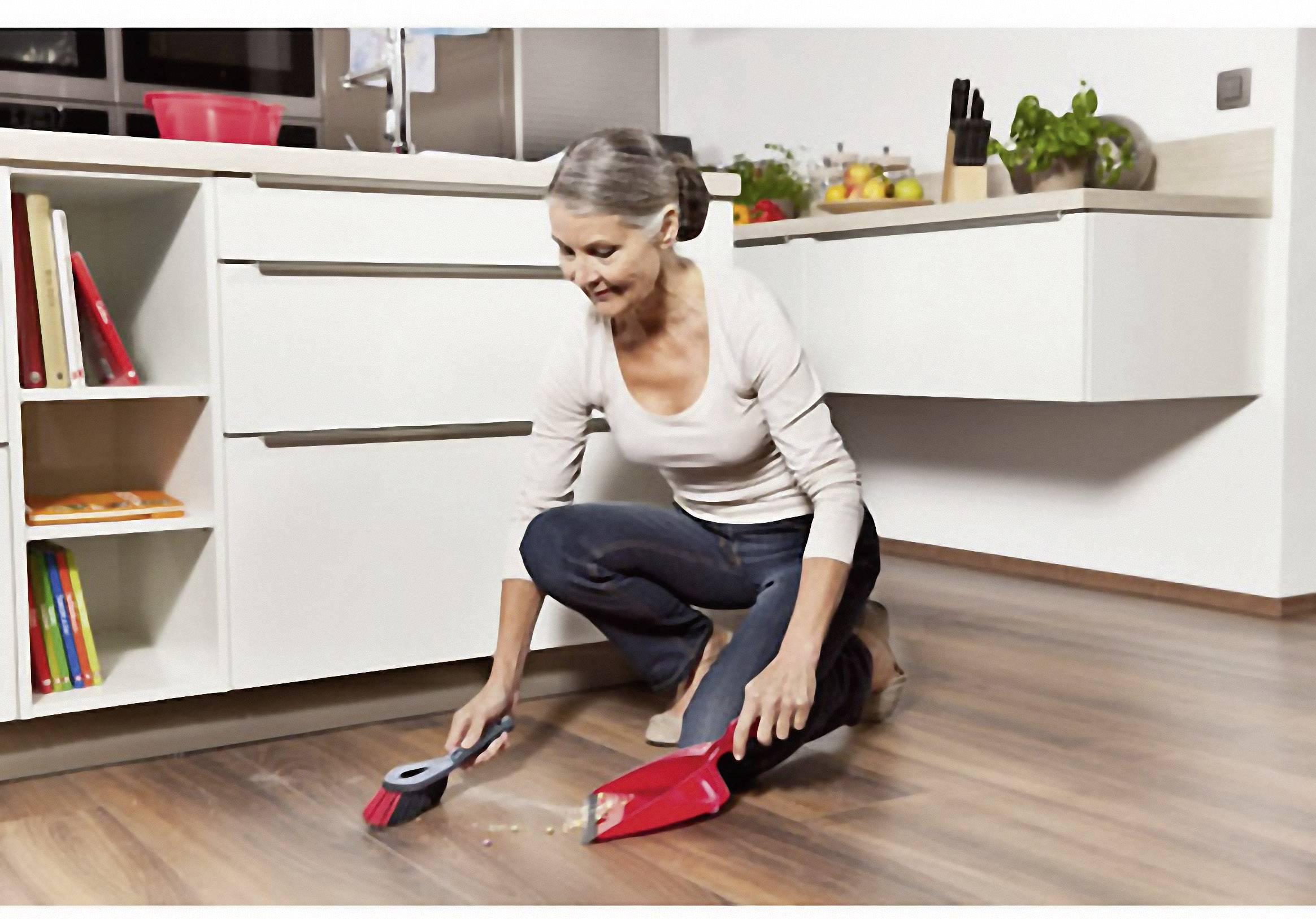 Une femme nettoie le sol d'une cuisine moderne avec un balai-brosse rouge et une pelle à poussière.