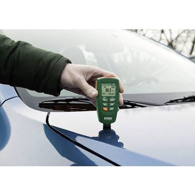 Une main tient un appareil de mesure d'épaisseur sur un toit de voiture bleu, affichant une épaisseur de 98,3 micromètres.