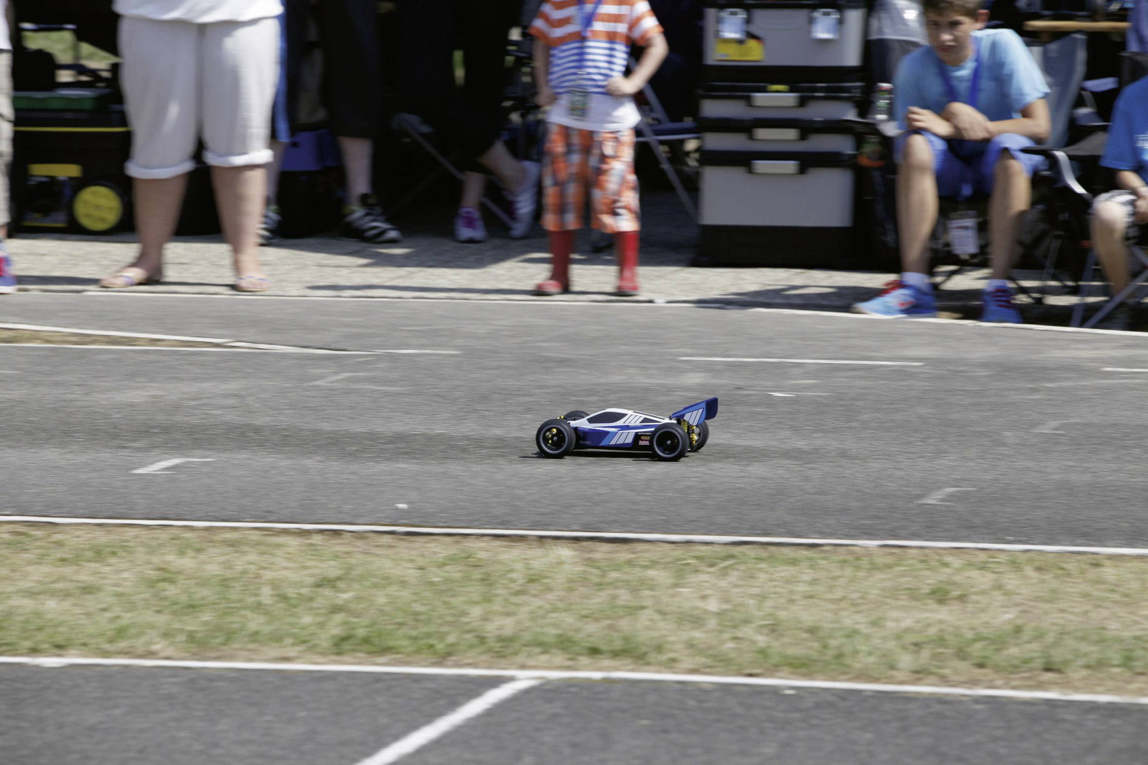 Une voiture télécommandée roule sur un circuit de course, entourée de personnes qui regardent. La voiture est noire et bleue.