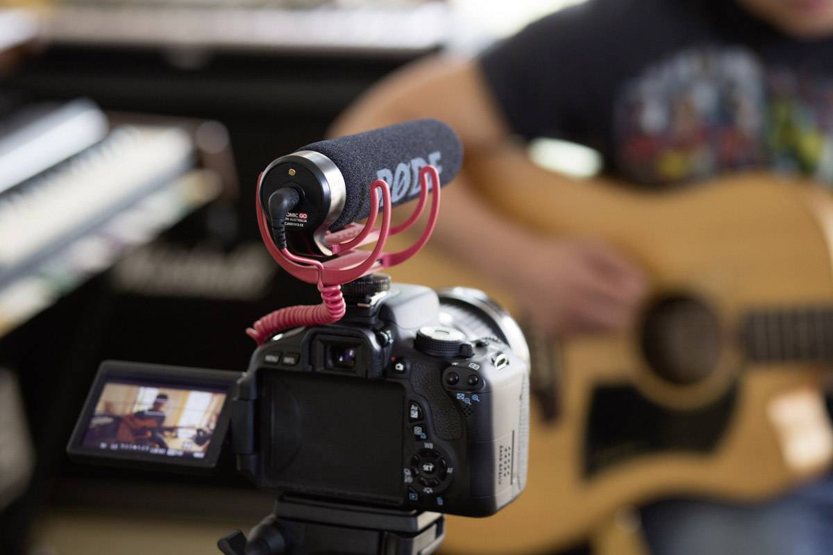 Une caméra équipée d'un microphone filme un guitariste. Un clavier et un ampli sont visibles en arrière-plan.