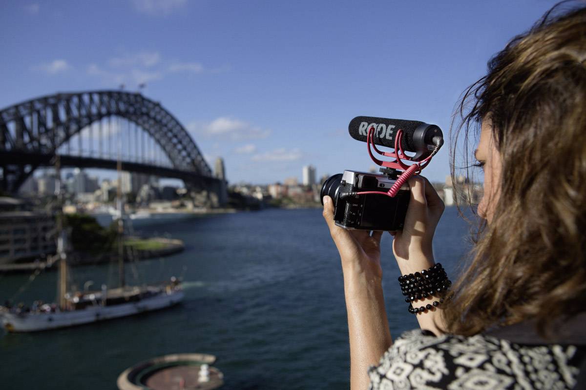 Une personne filme avec une caméra, le Harbour Bridge de Sydney en arrière-plan. Le temps est ensoleillé.