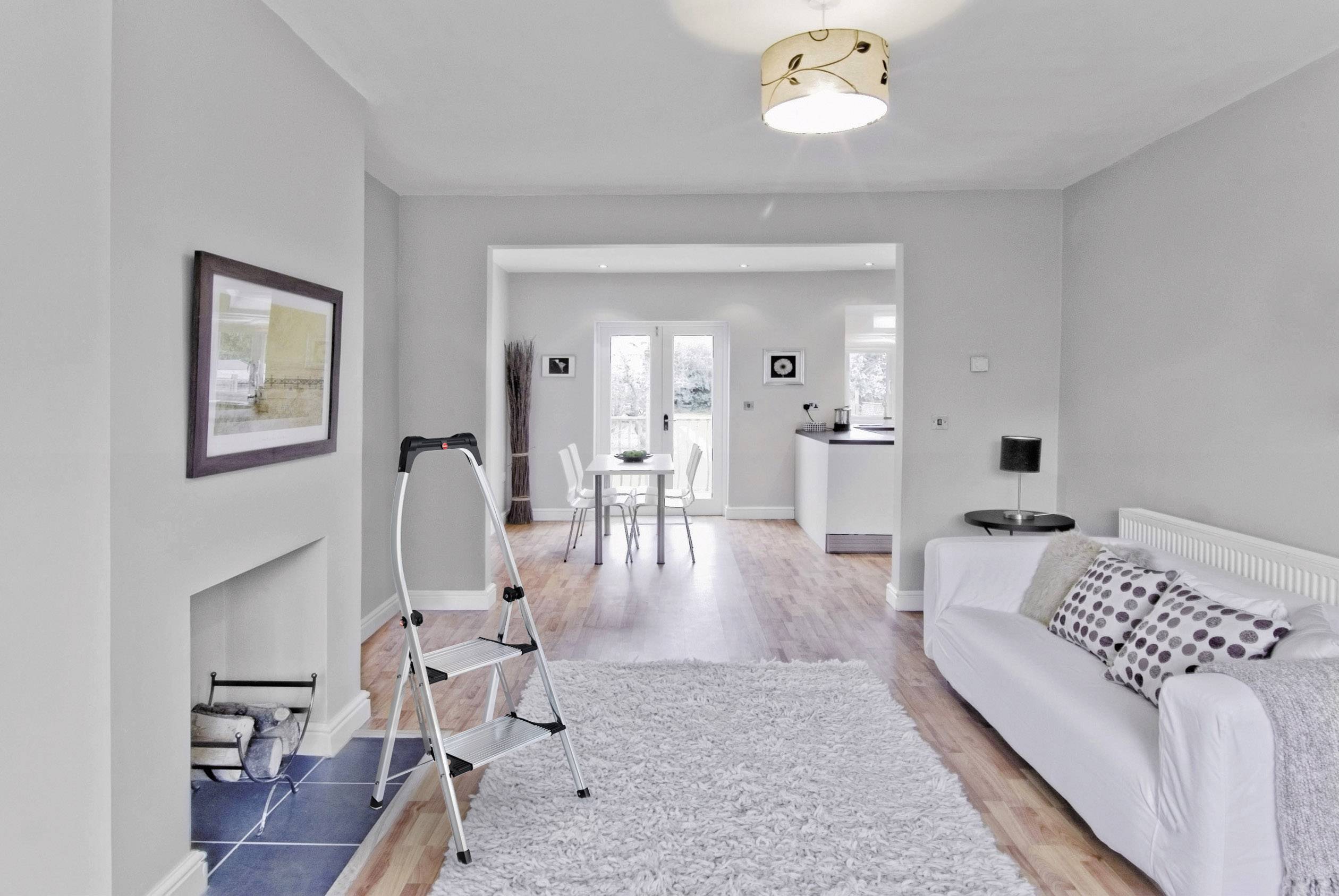 Salon lumineux aux murs gris, canapé blanc, échelle et tapis. Un coin repas et une cuisine ouverte sont visibles en arrière-plan.