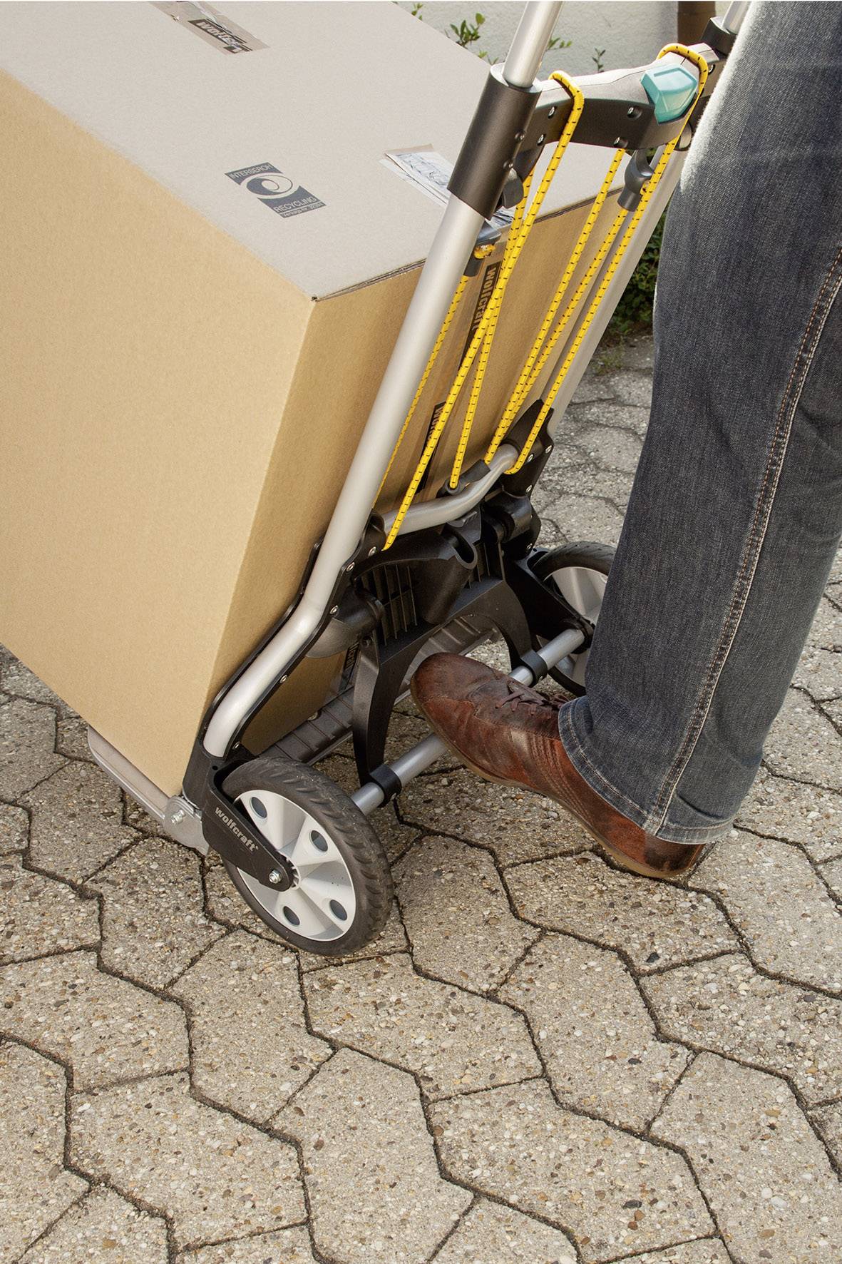 Une personne tire un chariot à main avec un grand colis sur un chemin pavé. La personne porte un jean bleu et des chaussures brunes.