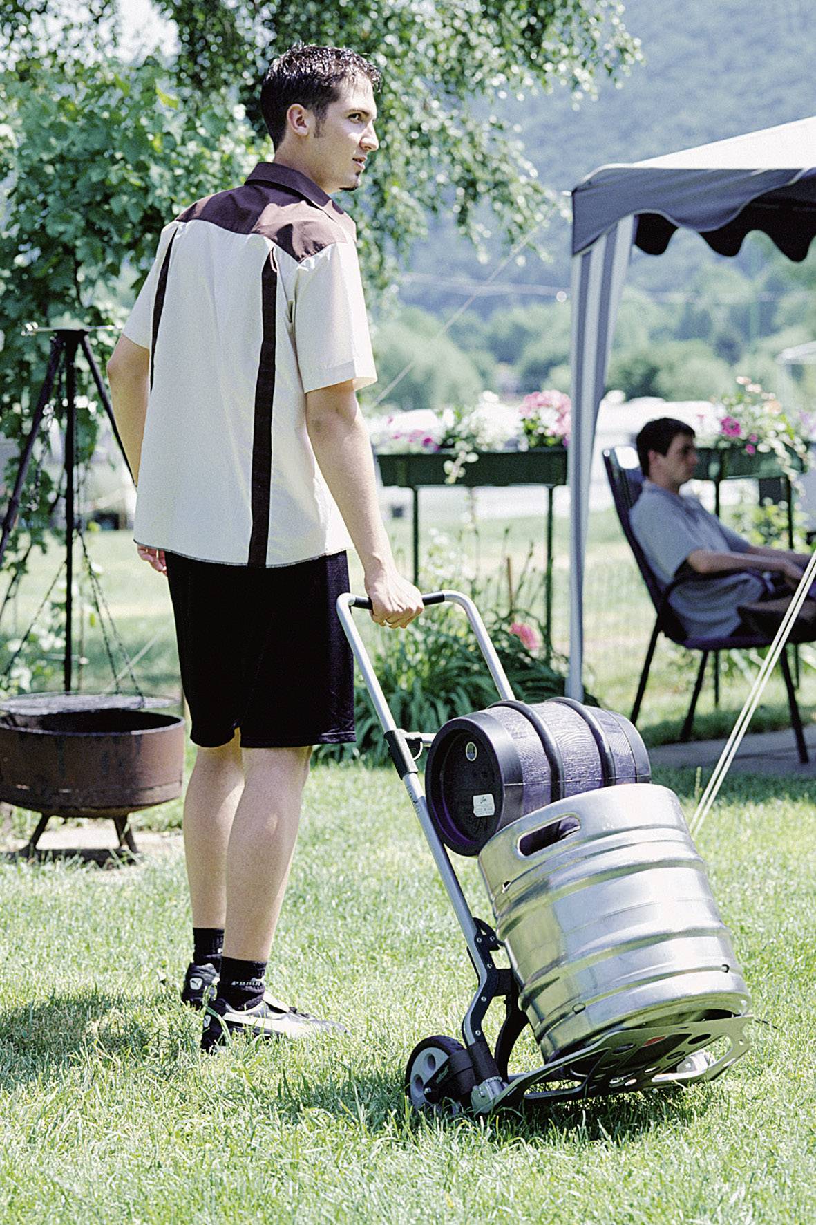 Une personne transporte des fûts de bière sur une charrette à bras par une journée ensoleillée dans un jardin, avec une tente et une personne assise en arrière-plan.