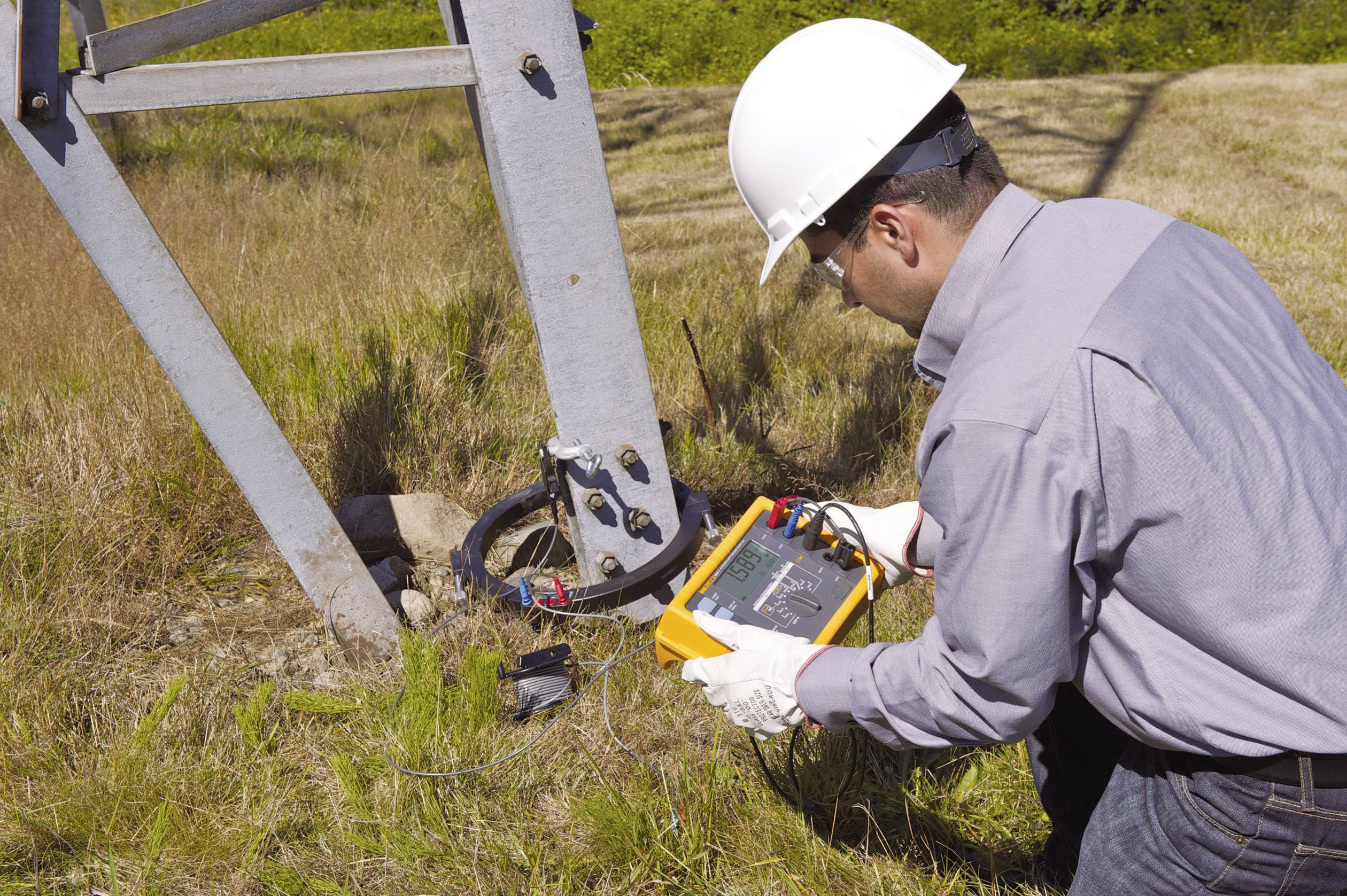 Un homme portant un casque de protection et des gants examine la base d'un mât en fer avec un appareil de mesure dans une prairie.