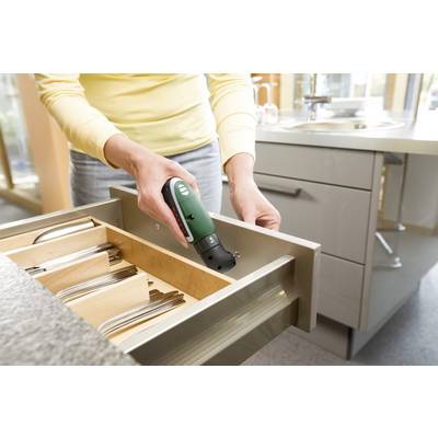 Une personne utilise une visseuse sans fil pour fixer un tiroir, entourée de meubles de cuisine. Des range-couverts sont visibles à l'intérieur du tiroir.