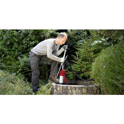 Une personne pompe de l'eau d'un puits à l'aide d'une pompe électrique rouge, entourée de végétation dense et de buissons.