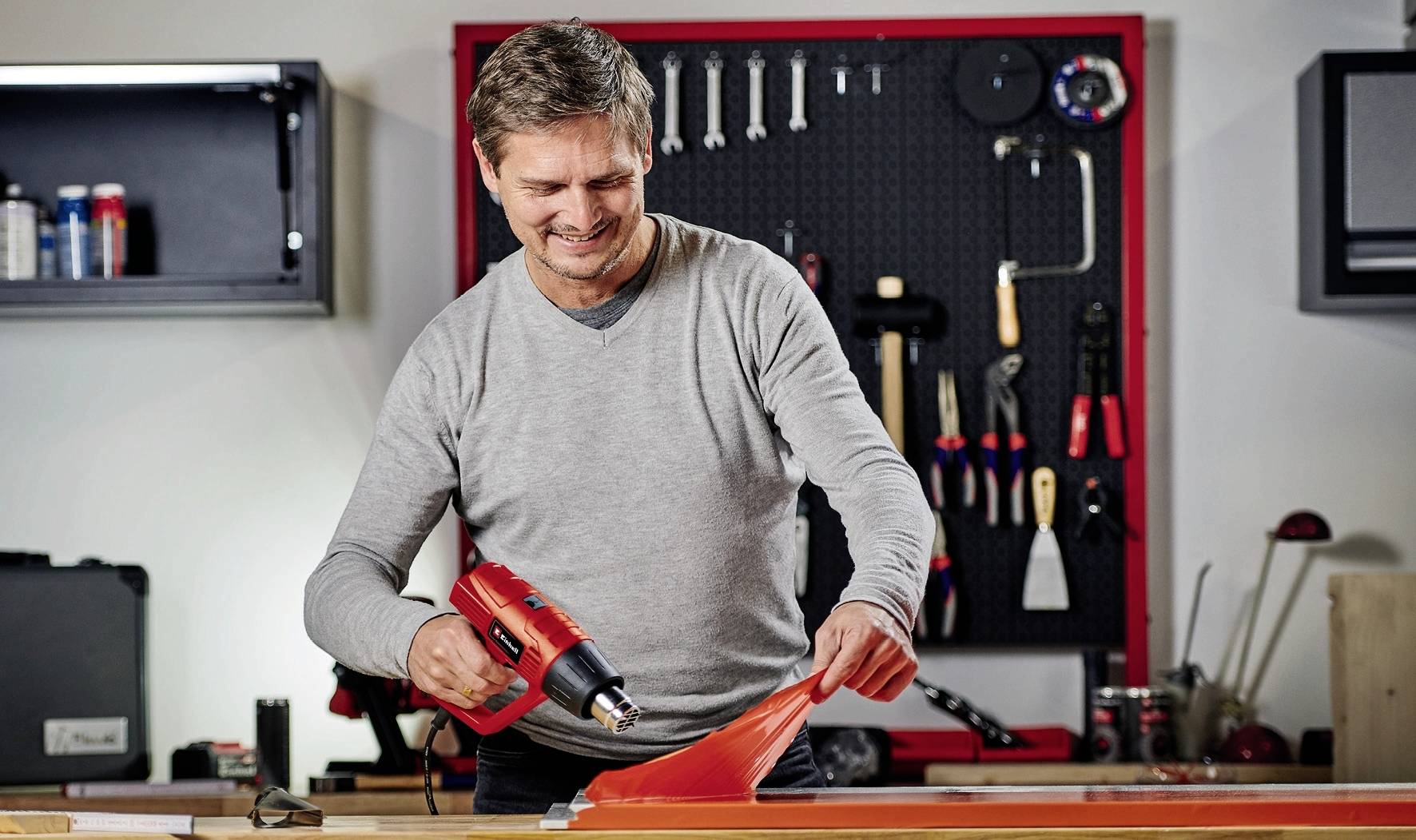 Un homme prépare un équipement de ski dans un atelier à l'aide d'un sèche-cheveux thermique. L'arrière-plan montre des outils accrochés au mur.
