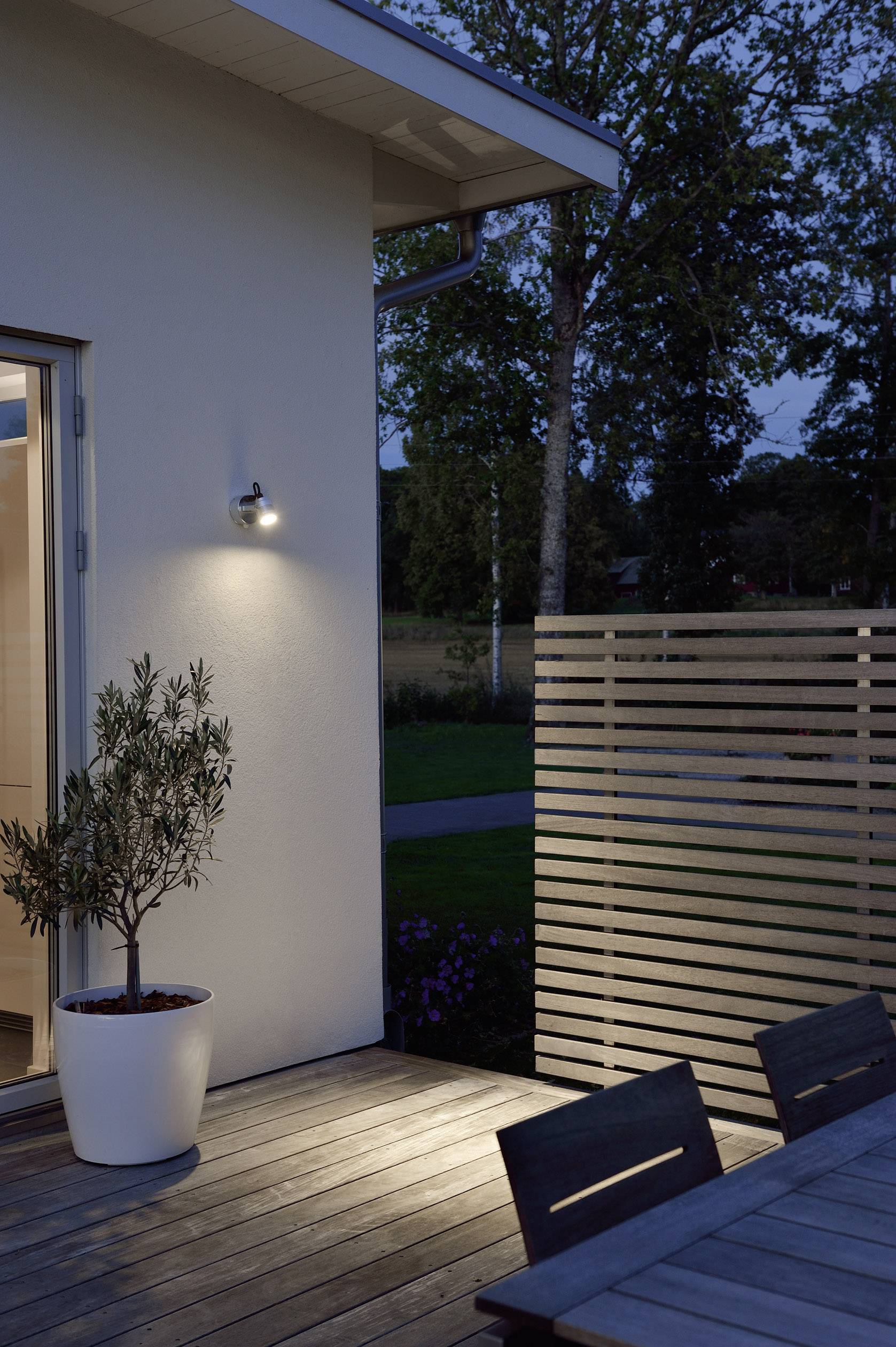 Une terrasse au crépuscule avec un espace extérieur éclairé. Un olivier en pot est placé à côté d'un mur en lattes de bois.