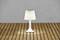Lampe de table blanche sur un plancher de terrasse en bois, illuminant une petite surface en dessous. Arrière-plan en béton gris.