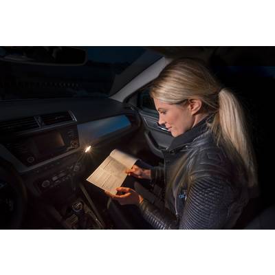 Une femme est assise dans une voiture dans l'obscurité et lit un manuel. Elle éclaire la page avec une petite lumière fixée dans la voiture.