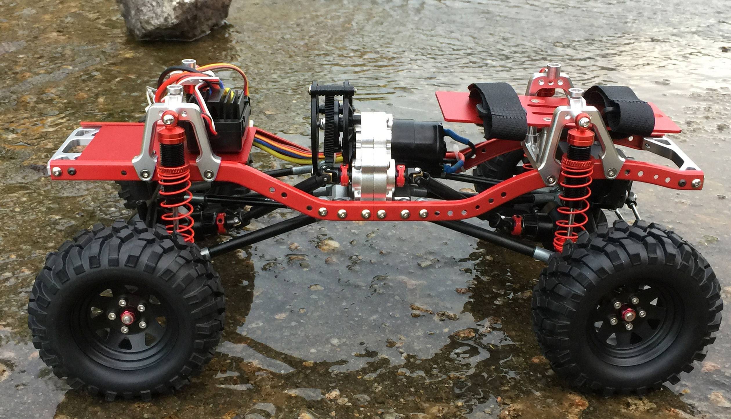 Un châssis de voiture tout-terrain télécommandée rouge, dépourvu de carrosserie, est posé sur un sol rocheux et humide. Il présente une suspension et des pneus détaillés.